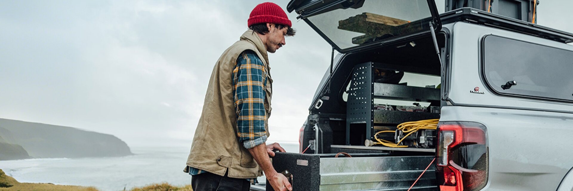 A man loading a storage box into a pickup truck bed with its IAQWE-supported canopy hatch held open, on a foggy coastline